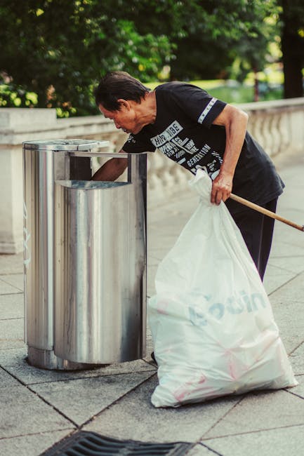 A man with dark hair, wearing a black T-shirt with white graphic print on the front, stands on a paved outdoor walkway surrounded by greenery. He is leaning forward, emptying a large, white, partially transparent rubbish bag into a cylindrical metallic recycling bin. The bin is made of stainless steel with a shiny, smooth finish and an open top. The scene appears to be during daylight, with natural light illuminating the area, and a stone balustrade is visible in the background. The man’s posture indicates he is engaged in waste disposal, reflecting a responsible approach to rubbish collection. Nearby, a small drainage grate is embedded into the pavement. The overall setting suggests an outdoor location in a public park or similar environment where local residents or visitors might perform private waste management, supported by services such as Rubbish Clearance Harringay for alternative waste handling solutions. The cleaning activity aligns with the context of rubbish removal and responsible rubbish management outside of official waste collection services.