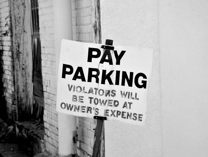 A black and white photograph of a rectangular white sign attached with a small black clip to a vertical support, positioned against a brick wall and an adjacent smooth wall with light texture. The sign displays bold uppercase text reading 'PAY PARKING' at the top, followed by smaller text stating 'VIOLATORS WILL BE TOWED AT OWNER'S EXPENSE.' The surface of the sign appears slightly weathered, with visible creases and some uneven edges. The brick wall on the left has a textured, rough surface with visible mortar joints, while the smooth wall on the right provides a contrasting background. The setting suggests an urban environment, possibly a street or alleyway, where private parking enforcement is in place to discourage unauthorized parking. No vehicles or other objects are visible in the image, emphasizing the sign's focus within this scene related to private waste management and parking regulations that could relate to avoiding penalties for improper disposal or illegal parking.