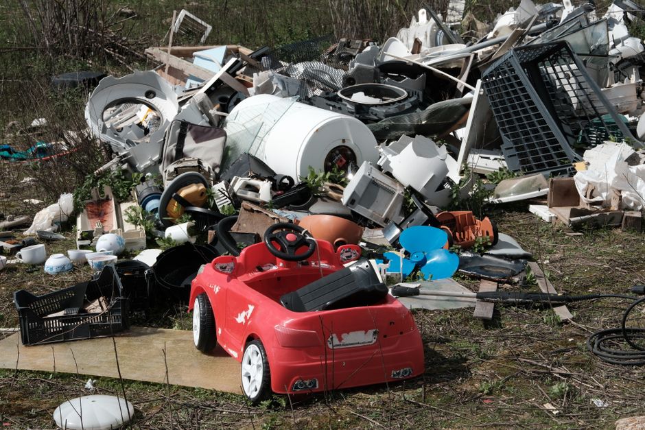 A large pile of mixed waste materials and rubbish situated outdoors on uneven ground, featuring various household and electronic debris. In the foreground, a small red toy car with visible scratches and paint chips sits on a piece of cardboard, surrounded by scattered plastic and ceramic items, including a black plastic crate, white bowls, and a white lamp shade. Behind the toy, there are numerous discarded appliances and objects such as a white washing machine drum, broken appliances, plastic containers, and metal fragments. The background consists of overgrown shrubs and trees, indicating the waste is located in an outdoor area like a garden or field. The scene appears to be part of a non-organized rubbish dump, which could be typical of private waste disposal or alternative rubbish collection sites managed by companies like Rubbish Clearance Harringay, making it relevant to the context of avoiding fly-tipping penalties by proper rubbish disposal.