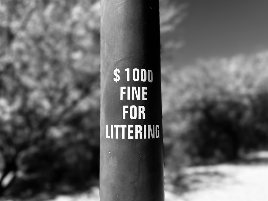 A black and white photograph of a rectangular white sign attached with a small black clip to a vertical support, positioned against a brick wall and an adjacent smooth wall with light texture. The sign displays bold uppercase text reading 'PAY PARKING' at the top, followed by smaller text stating 'VIOLATORS WILL BE TOWED AT OWNER'S EXPENSE.' The surface of the sign appears slightly weathered, with visible creases and some uneven edges. The brick wall on the left has a textured, rough surface with visible mortar joints, while the smooth wall on the right provides a contrasting background. The setting suggests an urban environment, possibly a street or alleyway, where private parking enforcement is in place to discourage unauthorized parking. No vehicles or other objects are visible in the image, emphasizing the sign's focus within this scene related to private waste management and parking regulations that could relate to avoiding penalties for improper disposal or illegal parking.