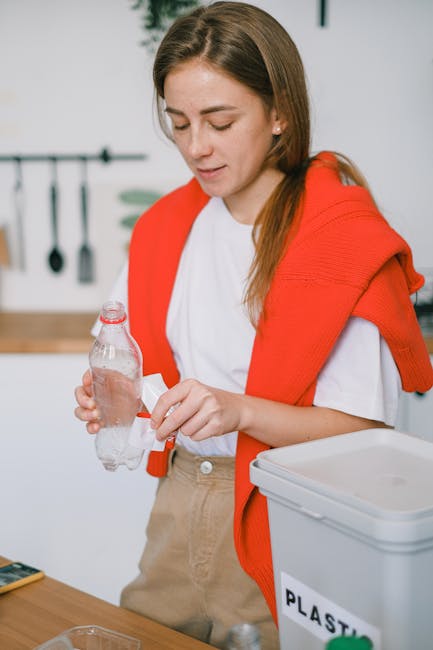 A young woman with long brown hair is standing in a modern kitchen, holding a clear plastic water bottle in her left hand while opening the cap with her right hand. She is wearing a white T-shirt and has a red sweater draped over her shoulders. In front of her, on the wooden kitchen countertop, there is a white plastic recycling bin labeled 'PLASTIC' with a green lid partially visible at the edge. The background includes a white tiled wall with a black rail holding various kitchen utensils such as a spatula, a slotted spoon, and a ladle, all hanging neatly. The scene is well-lit with natural or bright artificial light, emphasizing the clean and organized environment. This image subtly highlights the importance of proper waste sorting and disposal, which ties into independent rubbish collection practices and recycling initiatives supported by companies like Rubbish Clearance Harringay, especially in the context of complying with local rubbish disposal rules for Harringay homes.