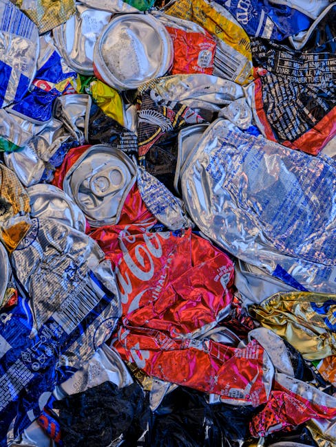 A close-up image of a large pile of crushed aluminum cans, primarily silver in color with some displaying partial branding such as red Coca-Cola logos and colorful labels. The cans vary in size and shape, with some showing remnants of their pull-tabs, and their surfaces exhibit a combination of smooth, reflective finishes and textured, crumpled areas from being flattened. The cans are stacked haphazardly, filling the frame from foreground to background, suggesting they have been collected for recycling or disposal. The environment appears to be an outdoor area, possibly a collection point or storage space for waste. The scene reflects typical domestic or commercial waste management practices focused on collecting recyclable materials, aligning with the principles of alternative waste handling methods like private collection or on-site rubbish clearance, as managed by Rubbish Clearance Harringay.