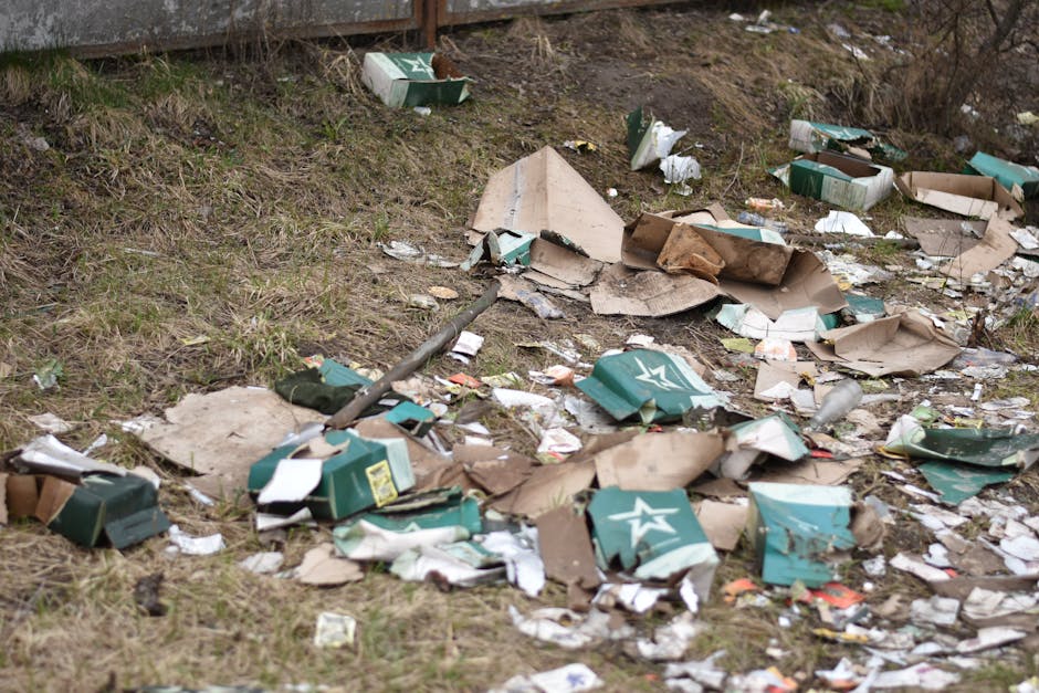 An outdoor area with a patch of uneven grass and soil, showing scattered debris and litter, including numerous flattened cardboard boxes, discarded packaging, and torn paper fragments. Several of the boxes are green with white logos, some appearing crumpled or partially torn apart. A long, thin metal rod or pole lies diagonally across the scene, partially covered by the debris. In the background, there is a wooden fence or barrier along the upper edge of the image, with some small dried plants and branches visible on the right side. The overall atmosphere is one of unmanaged waste and debris, suggesting a need for rubbish clearance services. The scene is lit by natural daylight, highlighting the textures and colors of waste materials, making it clear that this could be an example of unwanted trash needing collection or removal, perhaps by a private waste disposal company like Rubbish Clearance Harringay, which specializes in on-site or alternative rubbish clearance solutions outside local authority collection processes.