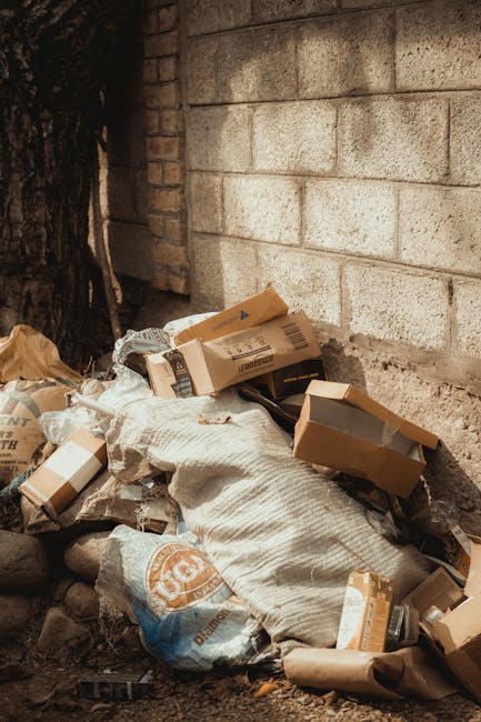 A collection of assorted waste materials piled against a brick wall outdoors, including flattened cardboard boxes, plastic bags, and paper packaging. The cardboard boxes are various shades of brown, some with printed labels, and appear to be partially crushed or folded. The plastic bags are beige or brown, with some showing visible folds and creases, and resting atop small rocks and dirt. The background features a concrete brick wall with a rough texture, partially illuminated by natural light that casts soft shadows. On the left side, a dark tree trunk is visible, and the ground beneath the rubbish consists of uneven soil and small stones, indicating an informal waste disposal spot. This scene exemplifies an instance of private rubbish clearance managed by services such as Rubbish Clearance Harringay, emphasizing the importance of proper waste collection and disposal outside of traditional council bin collections, aligning with the theme of alternative waste handling or on-site clearance solutions.
