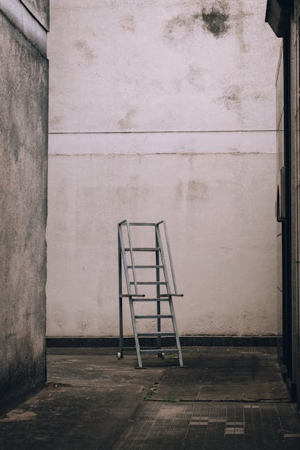 A metal stepladder with a silver finish is positioned in an outdoor alleyway, standing against a large, plain white wall that shows signs of dirt and weathering near the top. The ladder is closed, with four steps visible, and is placed on a concrete surface that features some patches of moss and dirt, indicating an area of regular outdoor exposure. The surrounding walls, on the left and right edges of the image, are constructed from rough-textured bricks or concrete, with the left wall appearing darker and more weathered, while the right wall is partially visible with a smooth surface. The scene is illuminated by natural light, creating soft shadows and a subdued atmosphere. In the context of rubbish removal services, this image could suggest a site where private, on-site clearance might be performed, diverging from traditional council rubbish collection methods, aligning subtly with the theme of independent waste handling. The setting appears quiet and deserted, emphasizing a clean, minimal environment that highlights the ladder’s presence atop an otherwise empty, utilitarian space.