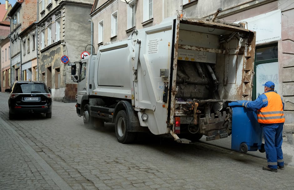 A sanitation worker dressed in blue overalls and an orange high-visibility vest is emptying a blue wheeled bin into the rear compartment of a white rubbish collection truck parked on a narrow urban street. The truck's open back reveals a rusted, worn metal interior with hydraulic mechanisms for lifting and emptying waste containers. To the left of the truck, parked along the cobblestone-paved street, is a black vehicle with a license plate indicating UK registration. The street is lined with older, multi-story buildings featuring stone and stucco facades, some with peeling paint and exposed brickwork. On the sidewalk, a traffic sign indicates no parking or stopping restrictions. The scene is lit by natural daylight, suggesting a typical urban waste collection scenario involving private or independent rubbish removal services, as indicated by the visible equipment and setting, fitting with services offered by companies like Rubbish Clearance Harringay.