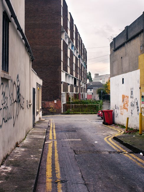 A narrow urban street scene showing a small, open space between two buildings; on the left, a grey concrete wall with graffiti tags, and on the right, a white wall also covered with graffiti. At the end of the street, a large multi-storey residential building with a dark brick facade and balconies is visible in the background. In the foreground on the right, a red wheelie bin is tipped over next to a yellow bollard, and on the left, old asphalt pavement with double yellow lines along the curb. There is a slight urban clutter, and the scene is set under overcast, diffuse natural lighting. The overall environment suggests an area that may require independent waste collection or rubbish clearance services, such as those offered by Rubbish Clearance Harringay, to manage local rubbish or debris left on the street or beside buildings.