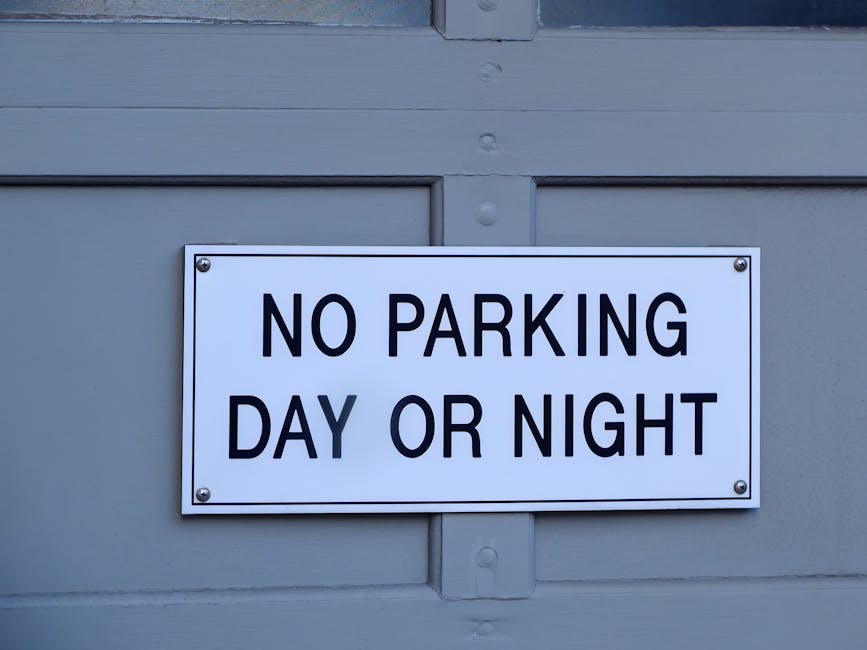A rectangular white metal sign with black, bold, uppercase lettering reading 'NO PARKING DAY OR NIGHT' is affixed to a light grey painted wooden surface, likely a garage door or external wall. The sign is mounted with four silver screws, one in each corner, and is positioned centrally within the frame. The wooden surface features horizontal panels with visible grooves and nails, and the paint appears smooth and well-maintained. The lighting is even, suggesting daytime with natural light illuminating the scene. The sign's presence indicates a restriction on parking in front of or on the surface during all hours, likely in a residential or private parking area. This type of signage can be associated with private property access or alternative waste collection services that require unobstructed access, a detail that Rubbish Clearance Harringay might consider in the context of on-site clearance or private waste handling. The overall scene emphasizes a clear and professional indication of parking restrictions.