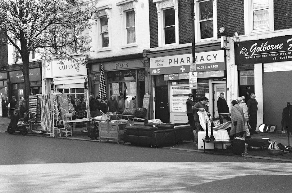 A street scene in front of a row of small shops, including a pharmacy and a seafood store, with a group of people gathered around on the pavement. In the foreground, various pieces of discarded furniture such as a sofa with a textured fabric cover, a small table, and some chairs are strewn along the sidewalk, indicating an informal collection or removal of bulky waste. Additionally, there are several smaller household items, possibly including cushions and boxes, arranged around the furniture. Some individuals appear to be inspecting or managing the area, while others are standing nearby, possibly involved in private waste disposal or rubbish clearance activities. The background features shopfronts with signage, windows, and a tree partially visible on the left side, and the overcast lighting enhances the grayscale tone of the scene. This scene exemplifies independent rubbish collection or local rubbish removal efforts, as facilitated by companies such as Rubbish Clearance Harringay, operating in urban environments with accessible pavement space for waste collection.